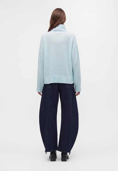Femme aux longs cheveux bruns portant un pull bleu clair, un pantalon large et ample sombre et des bottes noires, debout face à un mur blanc.