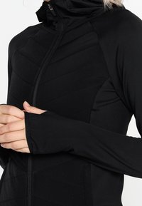 Black zip-up jacket with a high collar, featuring a textured, chevron pattern on the front and long sleeves with thumbholes.