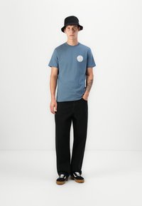 Blå bomulls t-skjorte med et rundt logo på brystet, kombinert med vide svarte bukser og svarte joggesko med hvite detaljer. Svart bøttehatt.