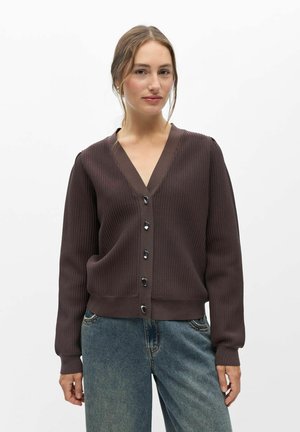 Femme portant un cardigan côtelé marron foncé avec des boutons et un jean bleu, debout devant un fond blanc uni.