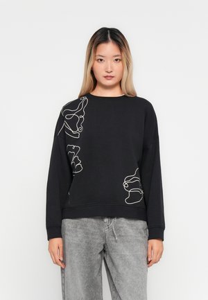 Kvinne iført en svart langermet sweatshirt med hvit abstrakt linjekunst og grå acid-wash jeans, som står mot en enkel hvit bakgrunn.
