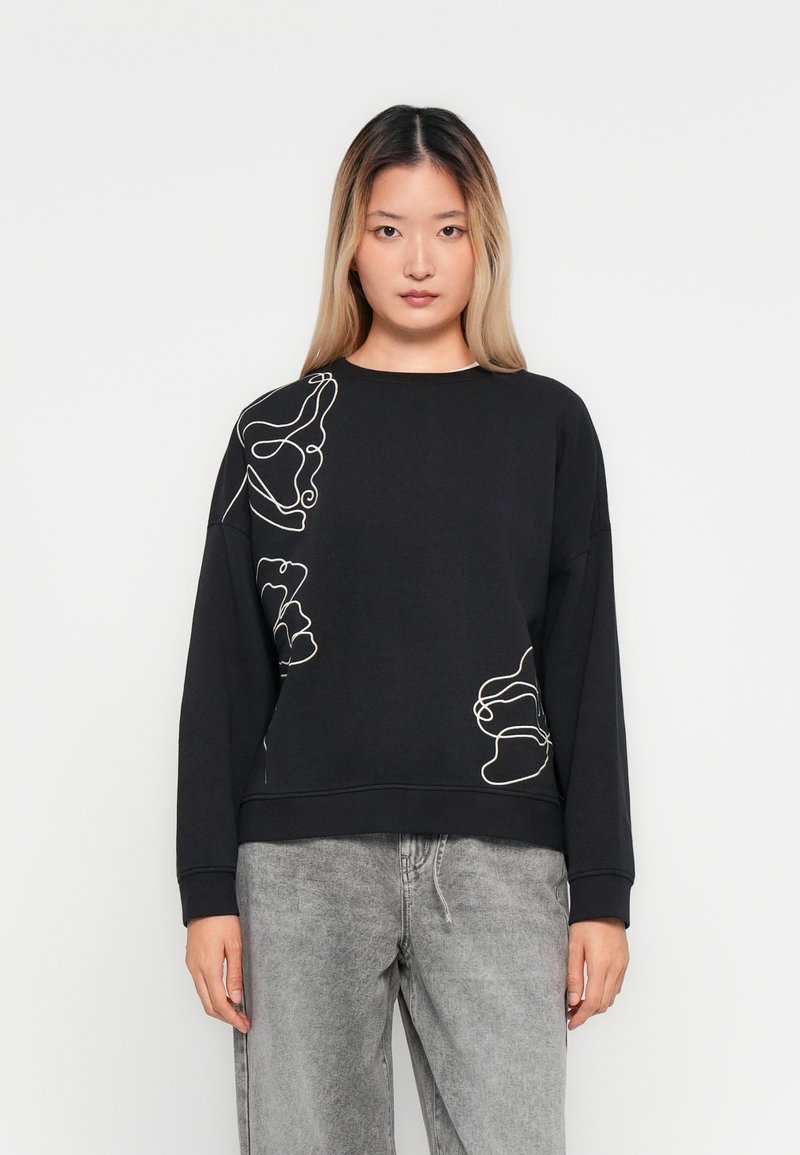 Kvinne iført en svart langermet sweatshirt med hvit abstrakt linjekunst og grå acid-wash jeans, som står mot en enkel hvit bakgrunn.