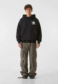 Fekete kapucnis pulóver elülső zsebbe és fehér grafikával. Olíva zöld cargo nadrág több zsebbel és logóval. Fekete cipők.