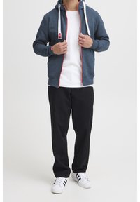 Uomo che indossa una felpa con zip blu navy con cordini bianchi, una maglietta bianca, pantaloni neri e sneakers bianche con strisce nere.