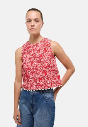 Femme portant un crop top rouge sans manches avec des motifs floraux blancs et un jean bleu taille haute, debout avec une main sur la hanche.