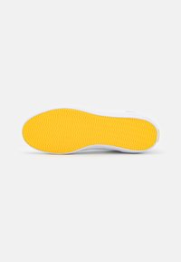 White athletic shoe sole with a bright yellow rubber bottom featuring a zigzag pattern for traction. Smooth texture and rounded edges.
