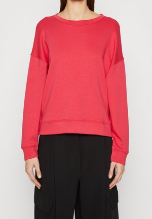 Person wearing a bright red long-sleeve crewneck sweatshirt and black pants with cargo pockets, standing against a plain background.