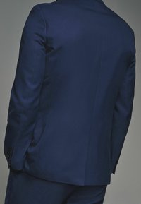 Granatowa, teksturowana marynarka o dopasowanym kroju, z jednym rozcięciem z tyłu i zapięciem na guziki przy mankietach. Szczegółowe przeszycia na ramionach.