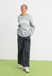 Grijze trui met "GODDESS" in wit en blauw, gecombineerd met zwarte bloemenpatroon wijde broek en witte sneakers.
