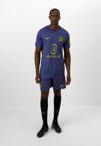 Lilla fotballdrakt med gula accenter. Innehåller logotyp, sponsortext och matchande shorts. Spelaren har svarta strumpor och fotbollsskor.