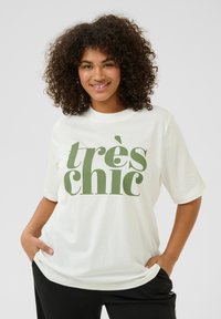 T-shirt en coton blanc avec une typographie verte surdimensionnée qui indique "très chic". Coupe décontractée avec des manches courtes et un col rond.