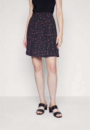 Frau trägt ein schwarzes ärmelloses Oberteil, einen schwarzen Rock mit kleinem rotem Blumenmuster und schwarze Sandalen mit Absatz, steht vor einem schlichten Hintergrund.