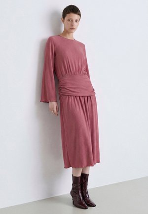 Donna con capelli corti che indossa un vestito midi rosa a maniche lunghe e stivaletti marrone scuro, in piedi contro un muro bianco semplice.