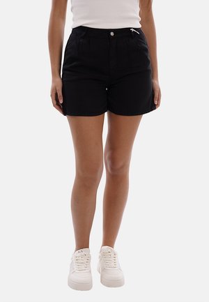 Donna che indossa shorts neri, top bianco senza maniche e sneakers bianche con lacci, in piedi davanti a uno sfondo chiaro uniforme.