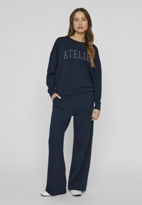 Tumši zilas krāsas džemperis un saskaņotas platās bikses, kas izgatavotas no mīkstas auduma. Džemperim priekšpusē ir izšuvumi ar vārdu "ATELIER".