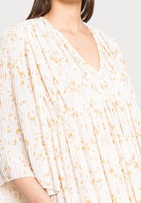 Femme portant une blouse blanche ample avec un délicat motif floral orange et un tissu plissé, dotée d'un col en V et de manches trois-quarts.