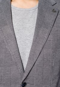 Szary i czarny marynarka w kratę z wyokrąglonym kołnierzem i jednolitym zapięciem na guzik, noszona na jasnoszarym T-shircie. Wzór zawiera różnorodne tekstury.