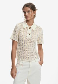 Pletený top v krémovej farbe s textúrovaným vzorom, s krátkymi rukávmi a kontrastným golierom, ktorý má dva hnedé gombíky. V kombinácii so svetlými nohavicami.