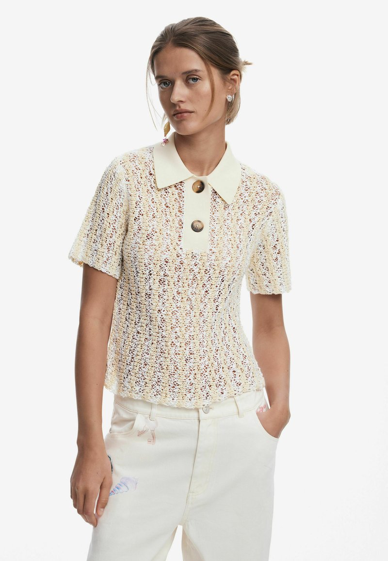 Pletený top v krémovej farbe s textúrovaným vzorom, s krátkymi rukávmi a kontrastným golierom, ktorý má dva hnedé gombíky. V kombinácii so svetlými nohavicami.