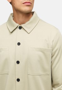 Lys beige knappjakke laget av strukturert stoff. Har en spredt krage, svarte knapper og to brystlommer.