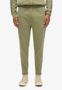 Olijfgroene joggers van zachte stof met zwarte zijstrepen, elastische tailleband en logo-detail. Gecombineerd met beige hoge sneakers.