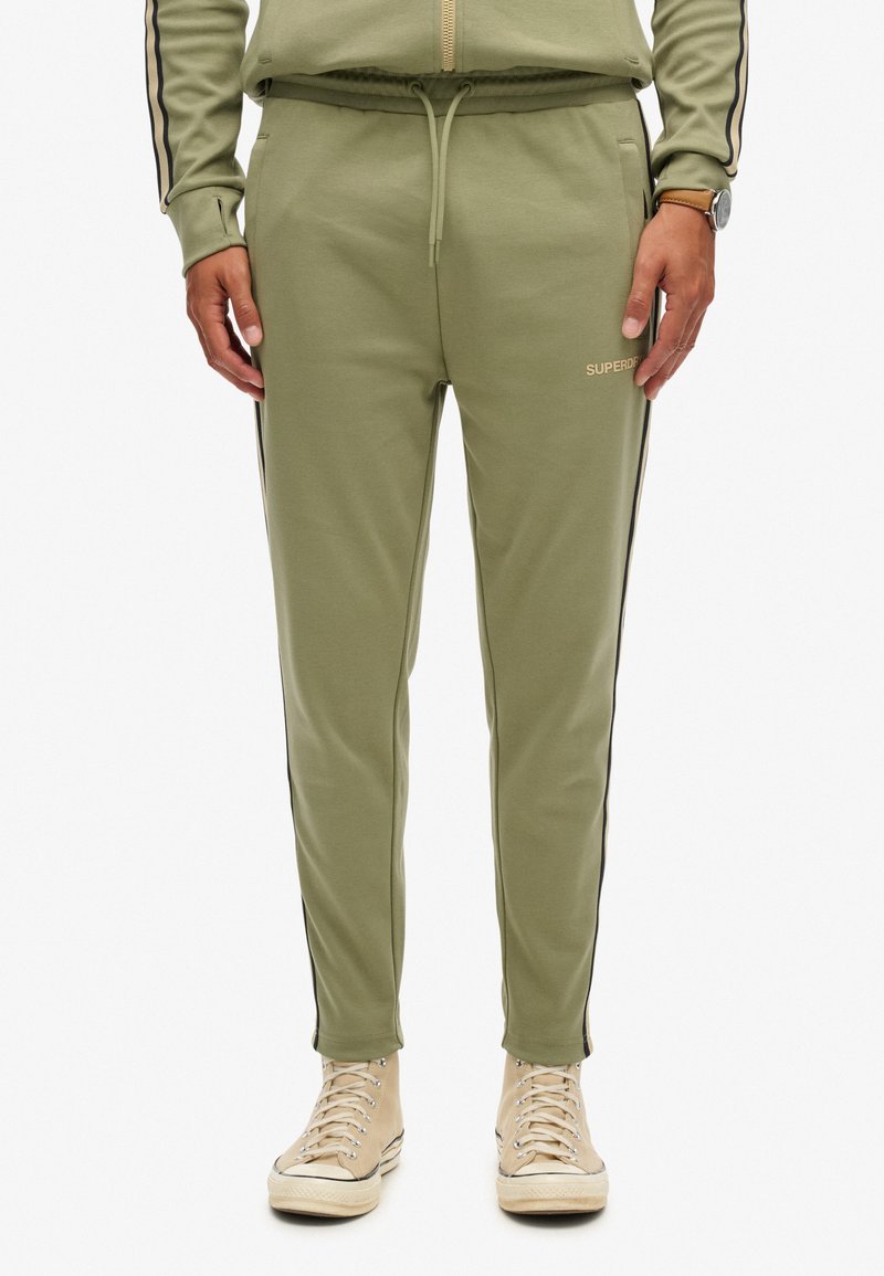 Olijfgroene joggers van zachte stof met zwarte zijstrepen, elastische tailleband en logo-detail. Gecombineerd met beige hoge sneakers.