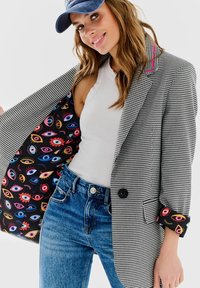 Sivá a čierna houndstooth sako s ružovými akcentmi, s farebnou podšívkou s očami, nosené cez biele tričko a modré džínsy.