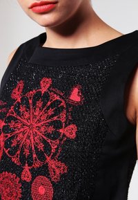 Närbild av en kvinna som bär en mönstrad svart ärmlös topp med röd intrikat cirkulär och hjärtformad broderi på framsidan.