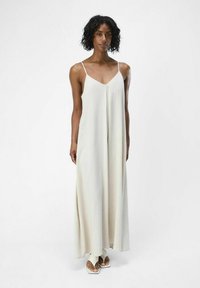Maxi dress color beige chiaro con spalline sottili e scollatura a V, caratterizzato da un design fluido e una sottile texture, abbinato a sandali semplici.