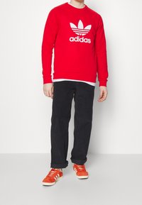 Röd sweatshirt med vit Adidas-logotyp, kombinerad med svarta byxor och orange sneakers. Sweatshirten har en rund halsringning och ribbade ärmslut.