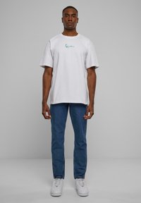 White short-sleeve t-shirt with green text logo on the chest, paired with blue straight-leg jeans and white sneakers. Simple design and fit.