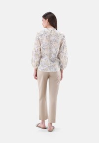 Floral blouse in sheer white fabric with yellow and gray patterns, puffed sleeves, and a high neckline, paired with beige slim-fit pants.
