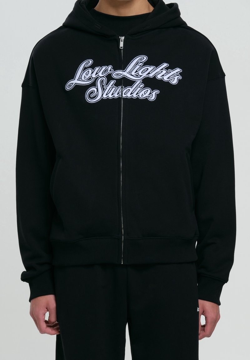 Personne portant un sweat à capuche zippé noir avec un texte blanc « Low Lights Studios » sur la poitrine, sur fond neutre.