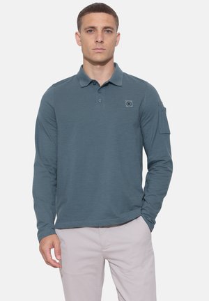 Jeune homme aux cheveux courts portant un polo bleu à manches longues et un pantalon clair, debout avec une main dans la poche.
