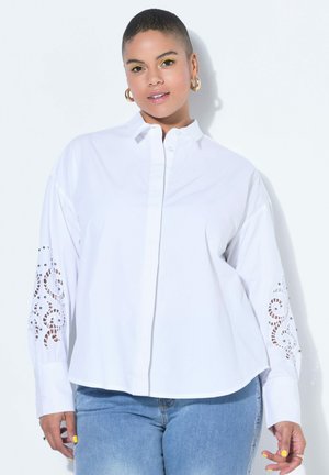 Frau mit kurzen Haaren trägt weiße Bluse mit ausgeschnittenen Mustern an den Ärmeln und blaue Jeans und posiert vor weißem Hintergrund.