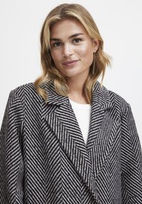Kabát s vzorom houndstooth v čierno-bielej farbe, s širokým reverom a veľkým golierom, so softílnou textúrou a štruktúrovaným strihom.