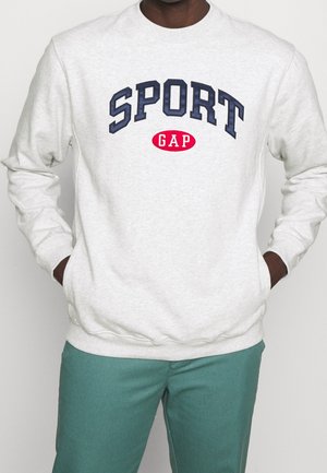 Osoba nosząca jasnoszary sweter z tekstem "SPORT GAP" w kolorach granatowym i czerwonym, ręce w kieszeniach, połączony z turkusowymi spodniami.