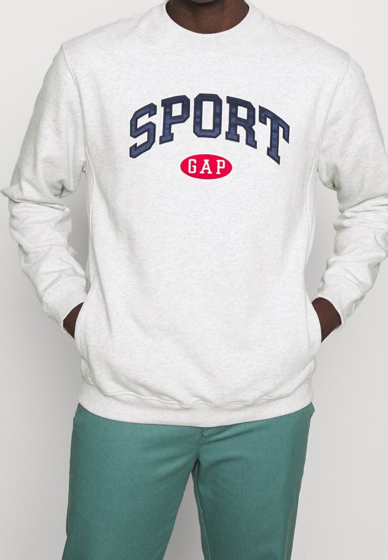 Personne portant un sweat-shirt gris clair avec le texte "SPORT GAP" en bleu marine et rouge, mains dans les poches, associé à un pantalon bleu sarcelle.