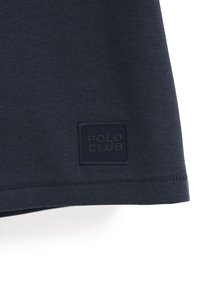 Camisa de manga corta de color azul marino con una textura suave. Presenta una etiqueta de tela cuadrada con "POLO CLUB" cosido en hilo oscuro en la parte inferior.