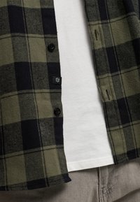 Chemise à carreaux verte et noire à boutons, portée ouverte sur un t-shirt blanc uni et un pantalon beige.