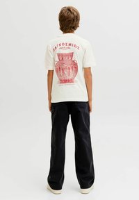 Crème t-shirt met een rode grafiek van een vaas en Griekse teksten op de achterkant. Gecombineerd met zwarte losse broek en lichtgekleurde schoenen.