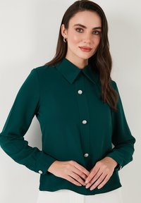 Dunkelgrünbluse mit Kragen, langen Ärmeln und dekorativen Edelsteinknöpfen entlang der Vorderseite. Glattes Material mit einem leicht schimmernden Finish.