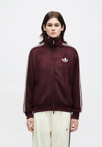 Jovem a usar uma blusa de treino com fecho éclair marrom-escura, com riscas brancas nas mangas, e calças de cor clara, contra um fundo liso.