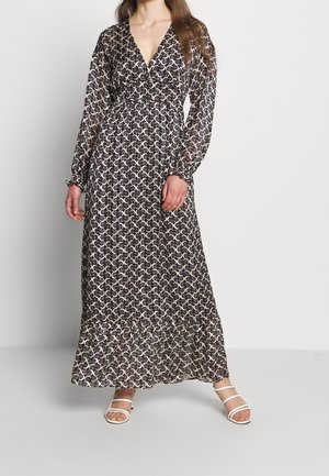 Kobieta ubrana w długą sukienkę maxi z dekoltem w serek, z wzorem czarno-biało-różowych zawijasów, oraz białe sandały na paski, stojąca na jednolitym tle.