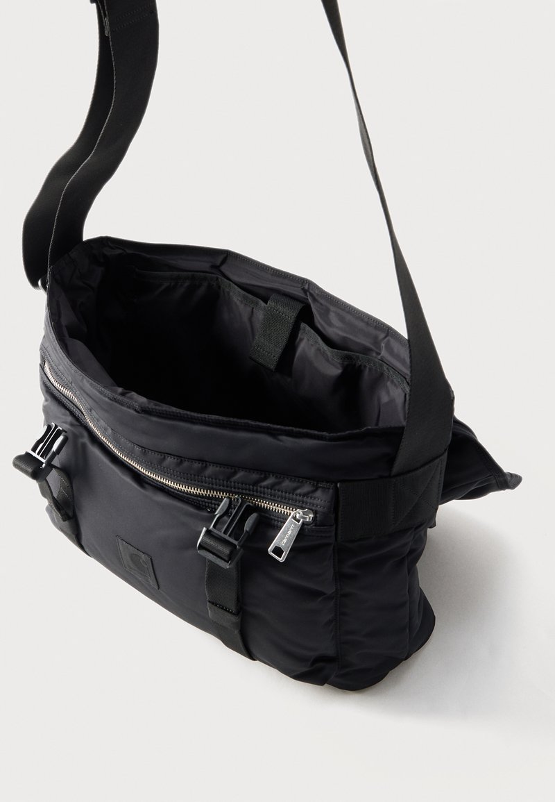 Sac bandoulière noir avec sangle réglable, ouverture zippée en haut, poche avant zippée et fermetures à boucle sur fond uni.