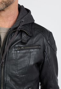 Zwarte leren jas met een ritszak op de borst, knoopsluiting bij de kraag en een grijze, gevoerde capuchon, voorzien van zichtbare stiksels en een gestructureerde afwerking.