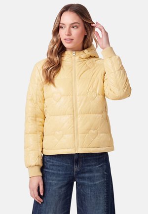Kvinde iført en lysegul dynejakke med hjerteformede quiltede mønstre, lynet op, kombineret med mørkeblå jeans, stående mod en ensfarvet baggrund.