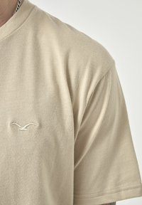 Beige Baumwoll-T-Shirt mit kurzen Ärmeln; verfügt über ein dezentes gesticktes Logo auf der Brust. Glatte Textur und entspannte Passform.