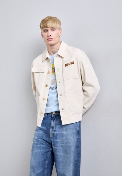 Blond man wearing a cream jacket with gold buttons over a light blue printed shirt and loose blue jeans, standing against a light gray wall.