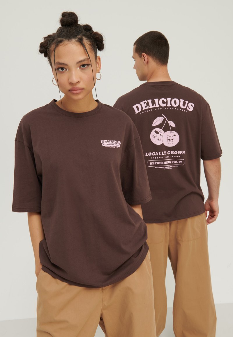 T-shirt oversize marron avec des impressions à l'avant et à l'arrière présentant des graphiques de cerises et du texte en rose. Tissu en coton doux, coupe décontractée, manches courtes.
